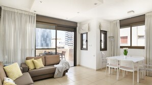 A modern living room with a sofa, a dining table with chairs, and large windows. - MAMAD Airy & Bright with Balcony & Parking steps from Beach by FeelHome (Tel Aviv - Jaffa)