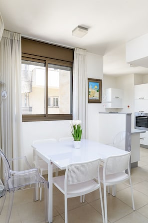 Uma mesa de jantar branca com cadeiras, uma janela com cortinas, uma área de cozinha e um quadro na parede.