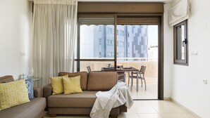A living room with a sofa, a glass table, and a view of a building through a large window. - MAMAD Airy & Bright with Balcony & Parking steps from Beach by FeelHome (Tel Aviv - Jaffa)