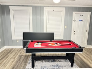 Une table de billard avec des billes et des queues de billard, placée devant un mur gris orné de deux portes blanches, dont l’une porte l’inscription « Interdiction de fumer ».