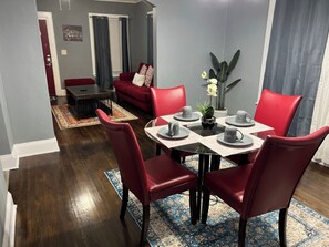 Un coin repas avec une table dressée pour quatre personnes, des chaises rouges, une petite plante en pot et, au fond, un canapé rouge.