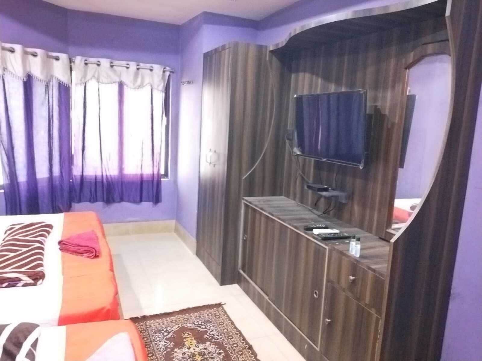 A bedroom with a bed, a TV mounted on a wooden cabinet, and a window with curtains.
