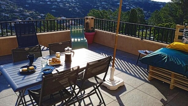 A balcony with a table, chairs, and an umbrella. There is a bed with a blue and yellow cover. The balcony overlooks a hilly landscape. - Appartement "rooftop" Avec Très Grande Terrasse à 2km de la mer (Roquebrune-sur-Argens)