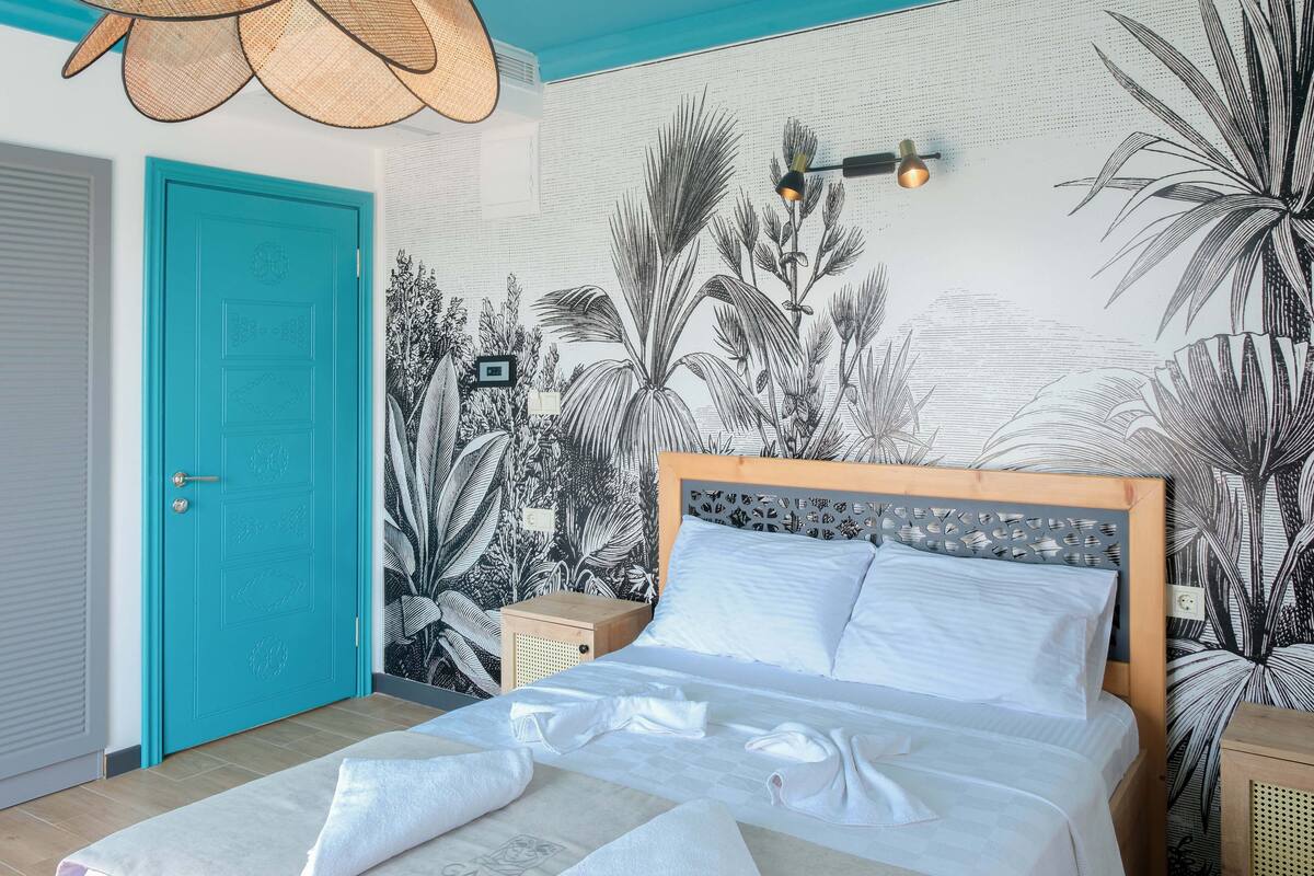 A bedroom with a teal ceiling, a bed with white linens, a wall mural of tropical plants, and a wooden door.