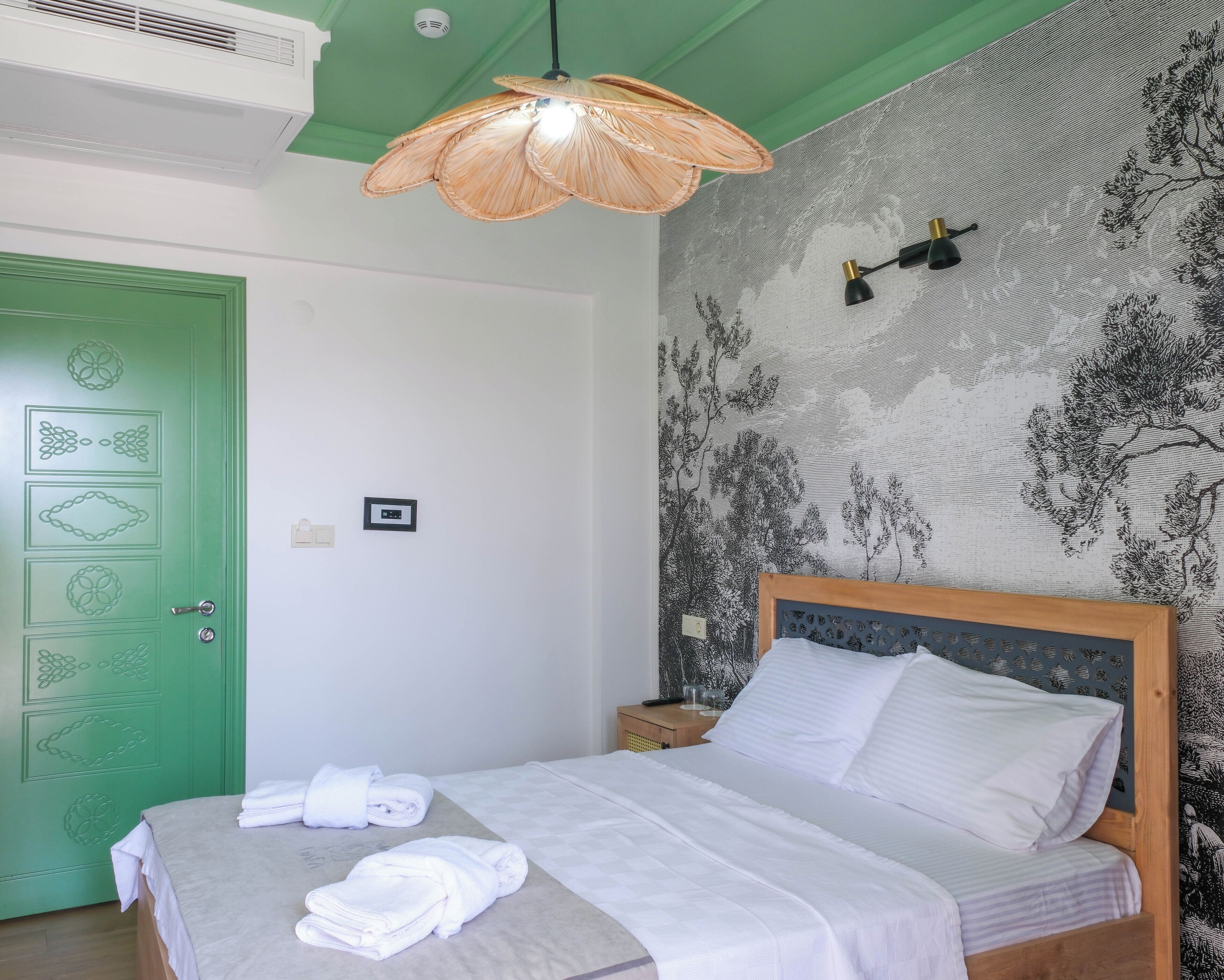 Una cama bien hecha con ropa de cama y toallas blancas, una puerta verde con diseños decorativos, un aire acondicionado de pared y una lámpara colgante.