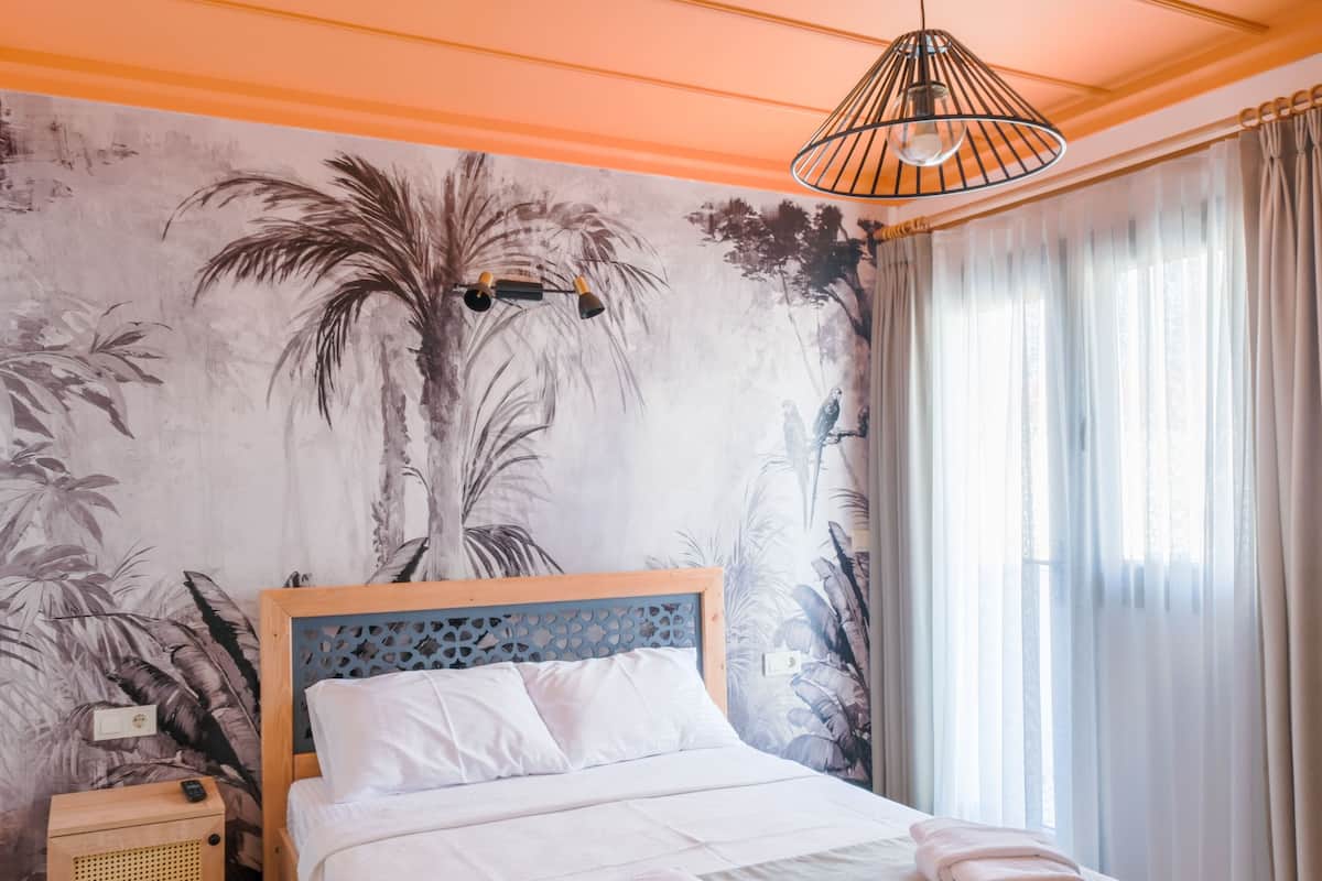 A neatly made bed with white linens and towels, a wooden nightstand with a speaker, a wall mural of tropical foliage, and a window with sheer curtains.