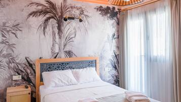 Una cama bien hecha con ropa blanca y toallas, una mesita de noche de madera con un altavoz, un mural de follaje tropical y una ventana con cortinas traslúcidas.