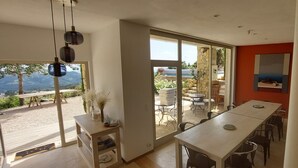Ein geräumiger Raum mit einer großen Schiebeglasdoor, die zu einer Außenpatiofläche führt. Im Inneren befindet sich ein langer Esstisch mit Stühlen, ein kleiner Beistelltisch und ein großer Spiegel an der Wand.