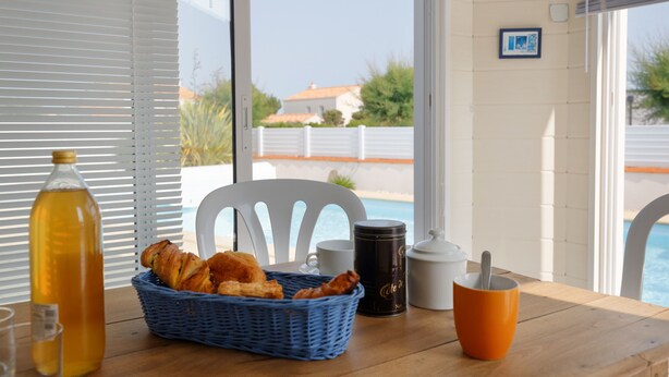 Una mesa de desayuno con un cesto de cruasanes, una botella de jugo de naranja, una taza de café, una taza negra y una tetera blanca.