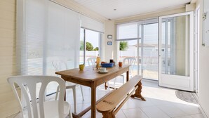 Un comedor con una mesa de madera, sillas blancas y un banco. Hay dos ventanas con persianas, una puerta y vistas al exterior.