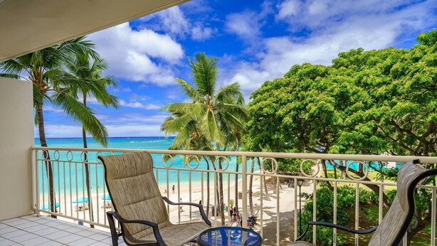 A balcony with two chairs and a small table, overlooking a beach with palm trees and a clear blue sky. - Ocean View Luxury Rental at Waikiki Shore #303! Steps to Beach! (Honolulu)