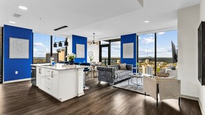 A modern kitchen with a central island, a dining area with a sofa and chairs, and large windows offering a city view.