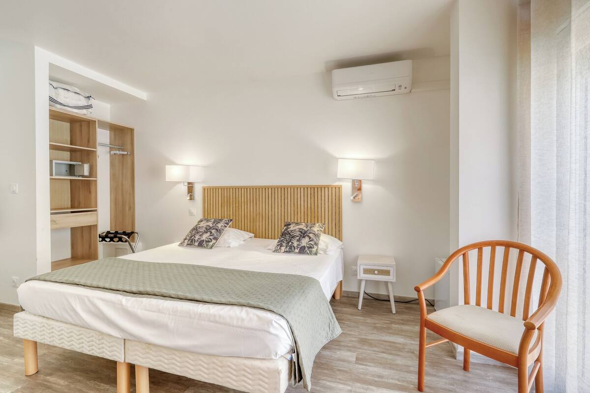 A bedroom with a bed, bedside tables, a chair, a wardrobe, and an air conditioning unit.