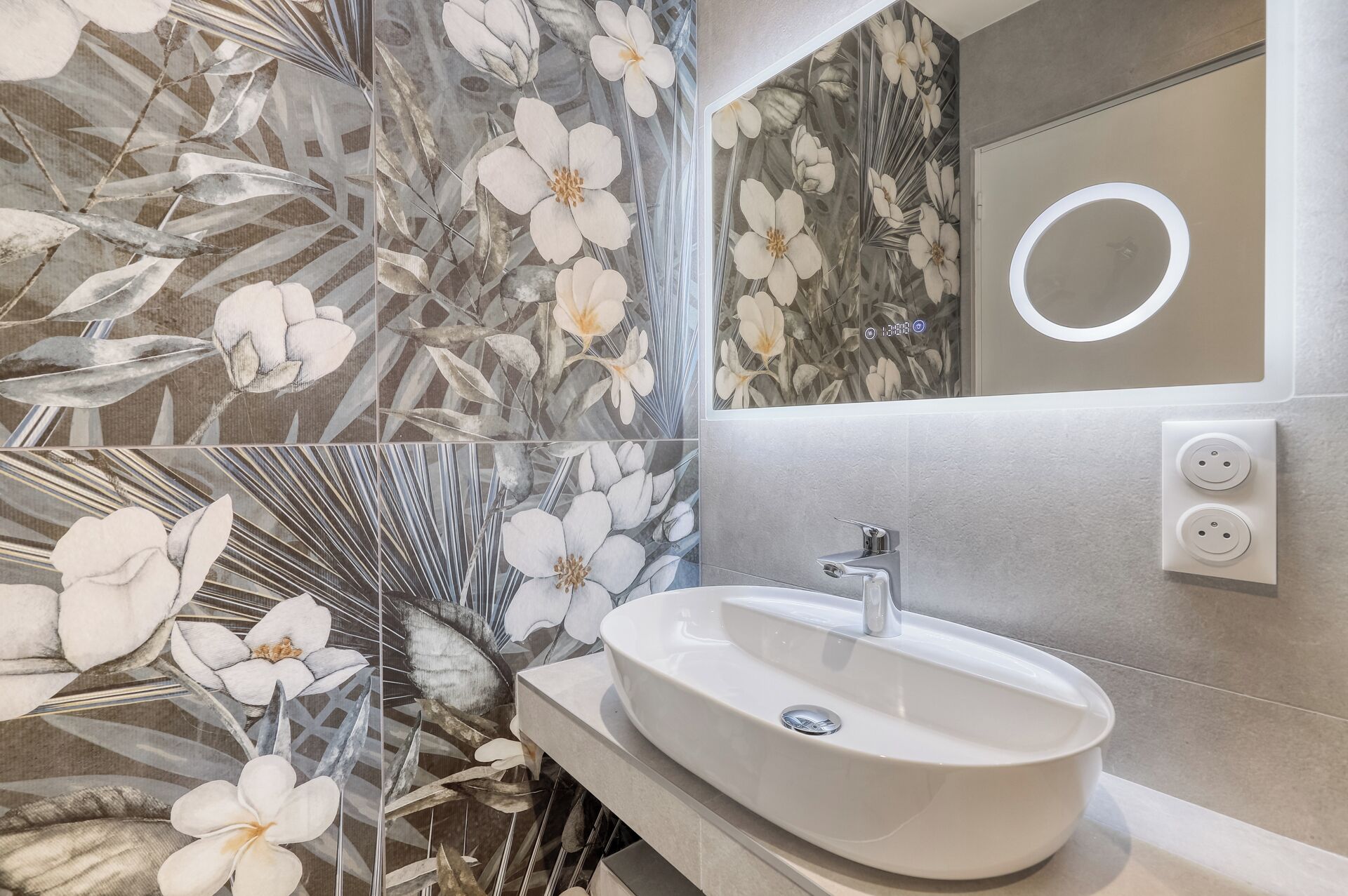 A bathroom with a white sink, a mirror with a circular light, and floral wallpaper.