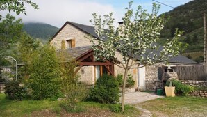A stone house with a wooden porch, surrounded by greenery and a stone wall.