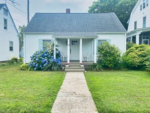 Une maison à deux étages avec un porche à l’avant, un petit jardin agrémenté d’hortensias bleus et une allée gravillonnée.
