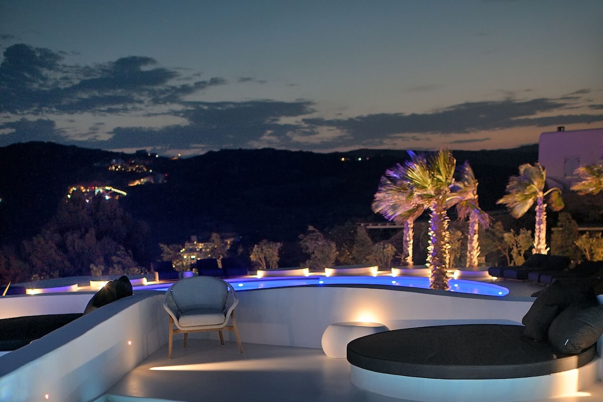 A modern infinity pool with built-in seating and ambient lighting, overlooking a scenic landscape with palm trees and distant lights.