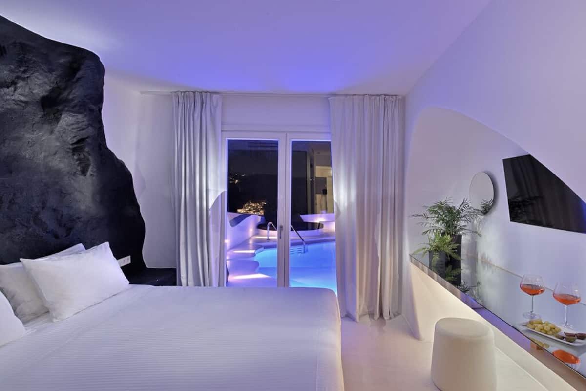 A modern hotel room with a large bed, a view of a pool, and a dining area with a mirror and table.