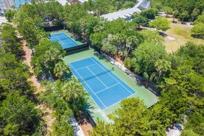 An aerial view of a tennis court surrounded by dense greenery and residential buildings.