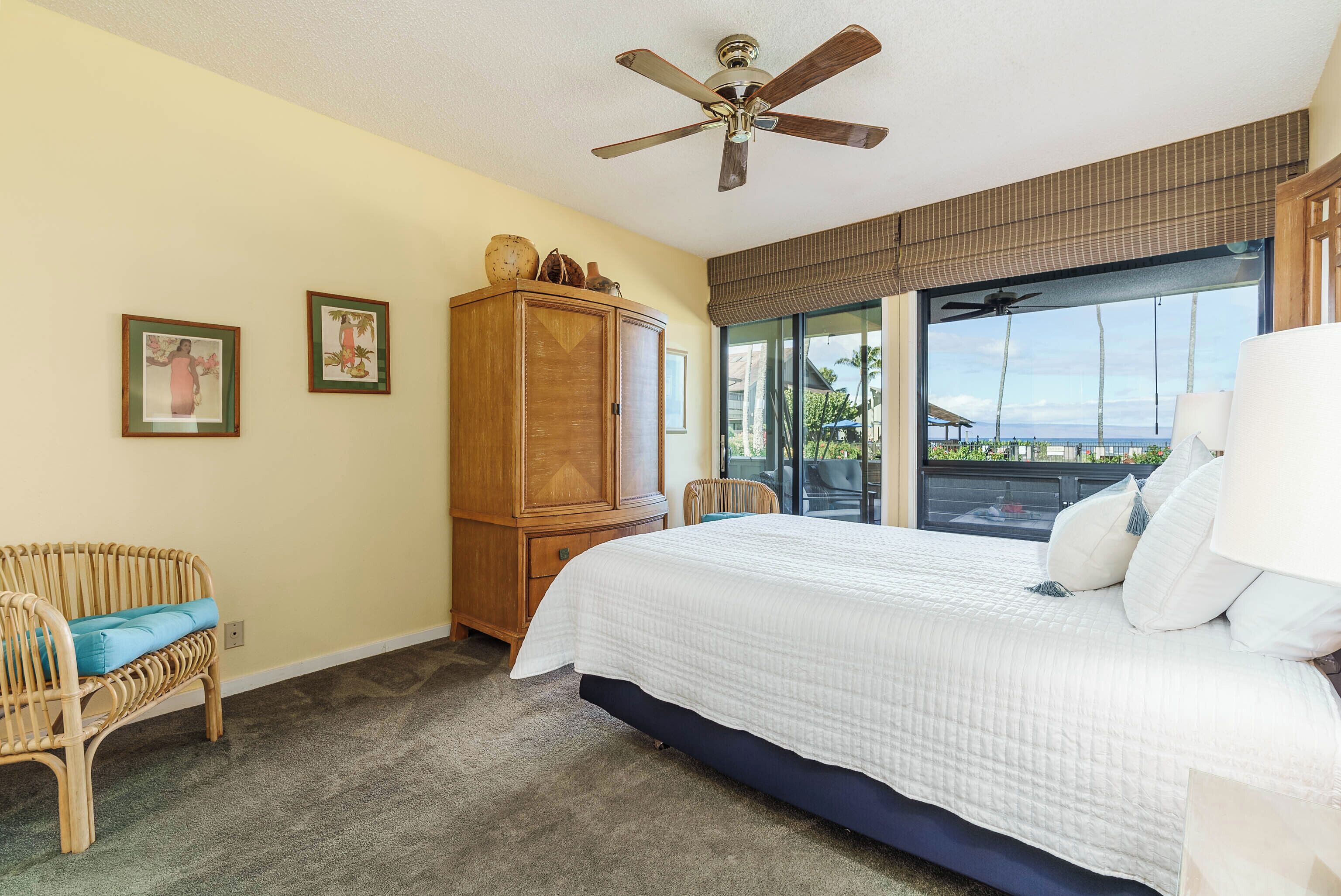 A bedroom with a large bed, a wooden wardrobe, a wicker chair, and a view of the outdoors through a window.