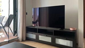 Una sala de estar moderna con un televisor de pantalla plana sobre un mueble negro, un área de comedor con mesa y sillas, y un puerta corrediza de vidrio que da a un espacio exterior.