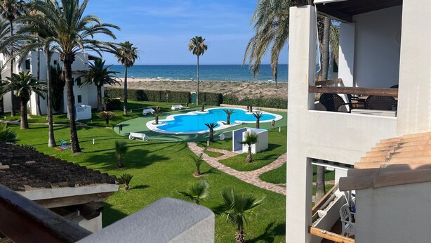 Zona de piscina con vista a la playa, rodeada de palmeras y un cielo despejado.