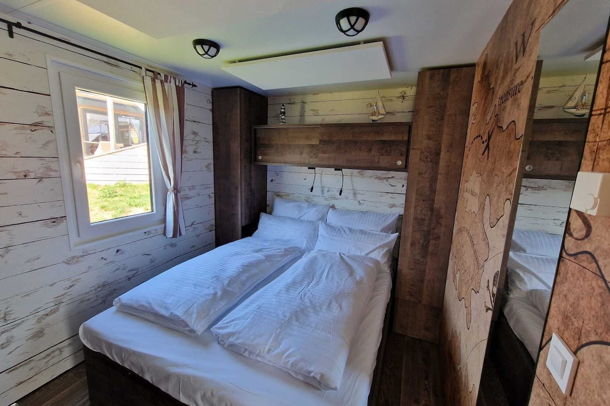 A bedroom with a wooden headboard, a large window with curtains, and a bed with white bedding.