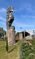 Una escultura de metal de un caballo sobre un pedestal de piedra en un área ajardinada, con un edificio al fondo.