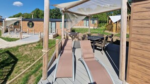 A wooden deck with outdoor seating and a shaded area, surrounded by wooden buildings with large windows. - Seaside-Resort (Geiselwind)