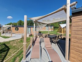 Una terraza de madera con mobiliario exterior y zona sombreada, rodeada de edificios de madera con grandes ventanales.