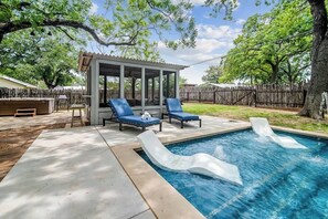 Un patio trasero con piscina, dos sillones de descanso, un área de estar cubierta y una valla de madera.
