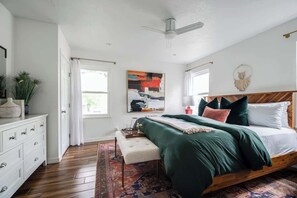 Un dormitorio con una cama de madera, un aparador blanco, un ventanal con cortinas blancas y un ventilador de techo.