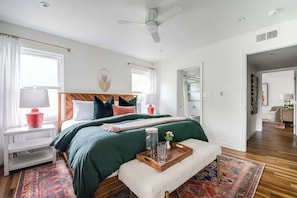 Un dormitorio con cama, mesitas de noche, una ventana, un ventilador de techo y una puerta que da a otra habitación.