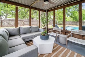 Un espacio exterior techado con un sofá modular gris, una mesita de centro blanca y una pequeña mesa redonda. Se ve una piscina al fondo.
