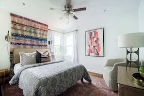 A bedroom with a bed, a ceiling fan, a window, a painting, and a lamp.