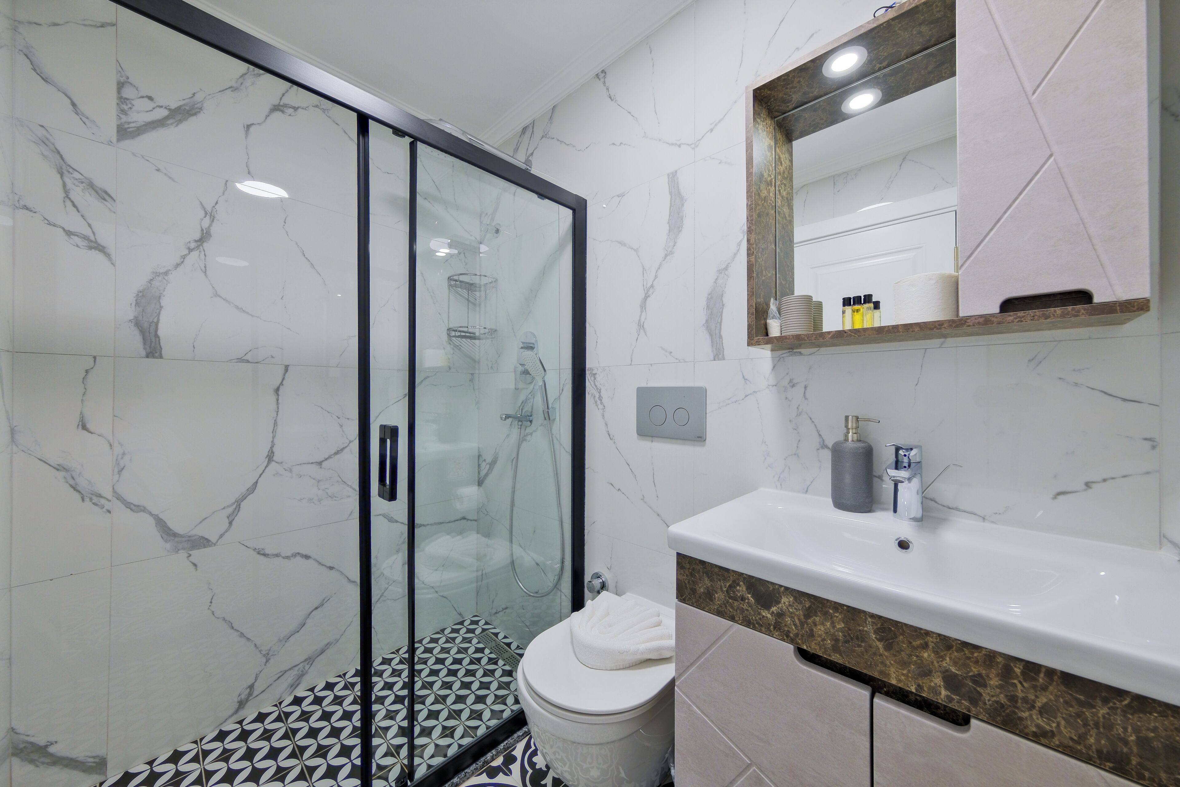 Un baño con cabina de ducha de vidrio, inodoro blanco, lavabo con encimera de mármol y espejo sobre el lavabo.