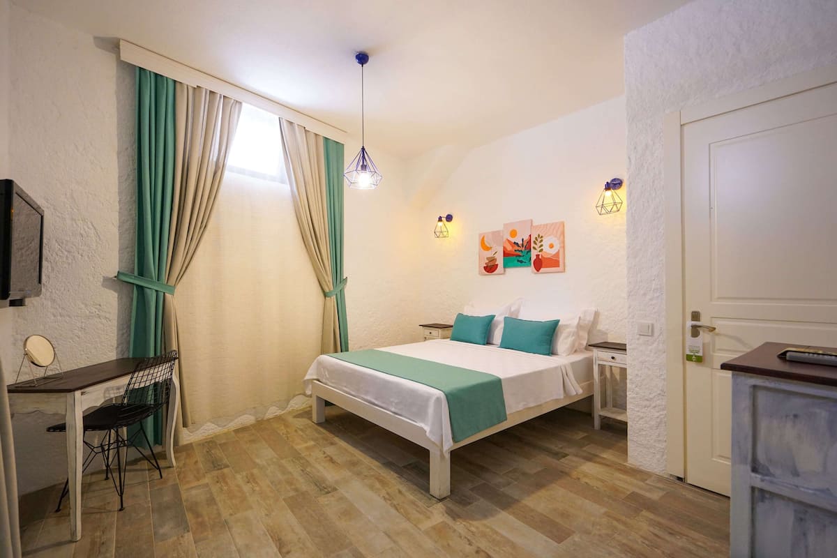 A bedroom with a bed, a desk, and a chair. There is a window with curtains, a wall-mounted light, and artwork on the wall.