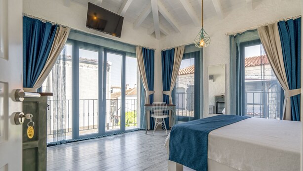 A bedroom with a bed, a desk, and a chair. There is a balcony with a view of buildings. - Portakal Cicegi Alaçatı  (Cesme)