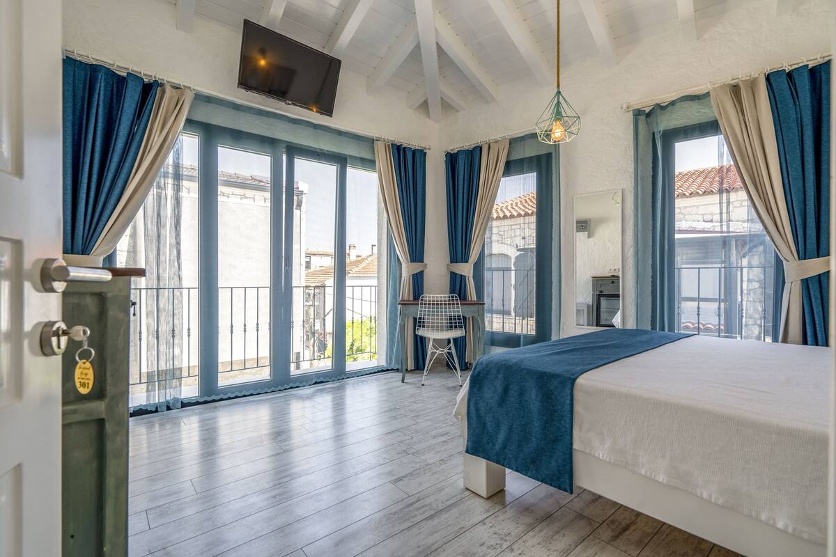 A bedroom with a bed, a desk, and a chair. There is a balcony with a view of buildings.