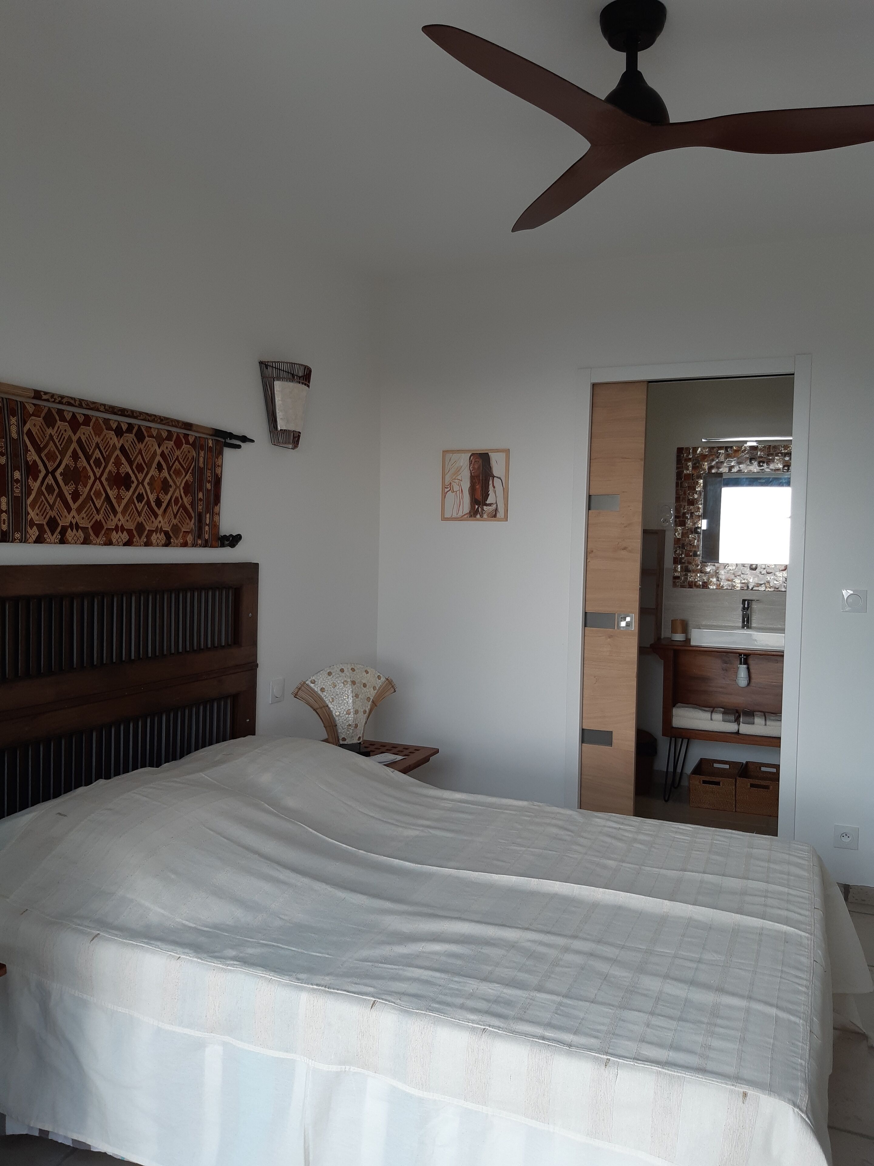 A bedroom with a bed, a ceiling fan, a wooden headboard, a wall-mounted shelf, a small table with a fan, and a doorway leading to a bathroom.