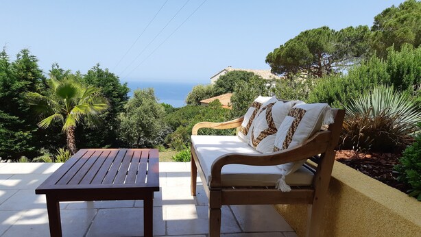 Une terrasse avec une table en bois et un banc rembourré, offrant une vue panoramique sur la mer et la verdure.