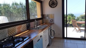 Une cuisine équipée d’une cuisinière à gaz, d’un évier et d’un lave-linge, avec de grandes fenêtres et une vue sur la verdure.