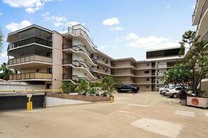 A multi-story residential building with balconies, a parking area, and potted plants.