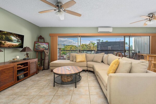 A living room with a sectional sofa, a round coffee table, a TV, and a large window with a view of palm trees and a building.