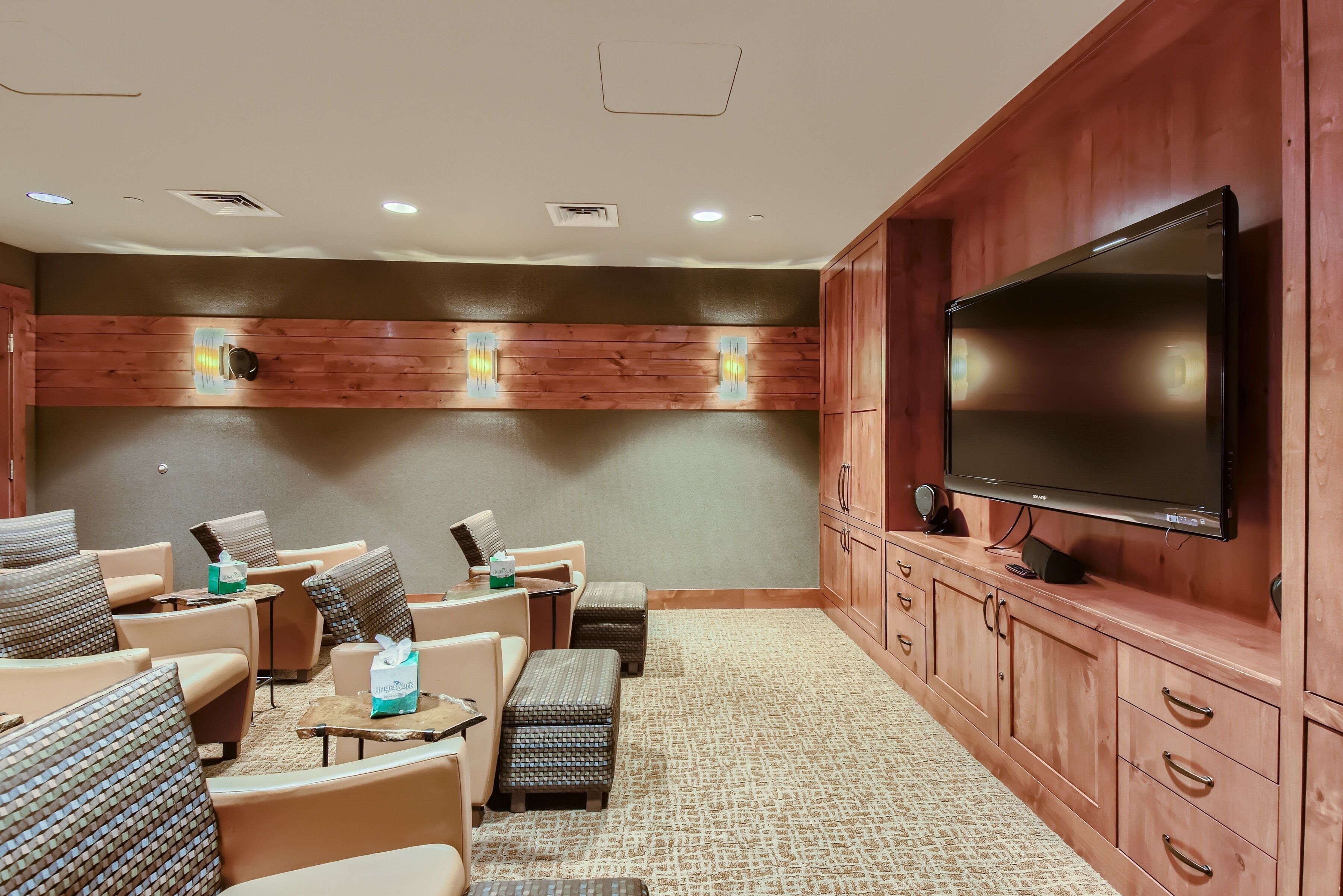A room with a large flat-screen TV, wooden panel walls, and multiple seating areas with cushioned chairs and ottomans.