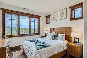 A bedroom with a large window, wooden headboard, bedside tables with lamps, and a view of a forest.