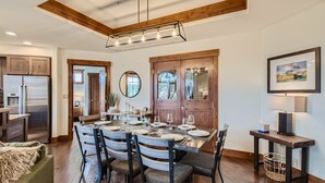 Un coin repas avec une table en bois dressée pour six personnes, entourée de chaises, d’un lustre et offrant une vue sur la cuisine.