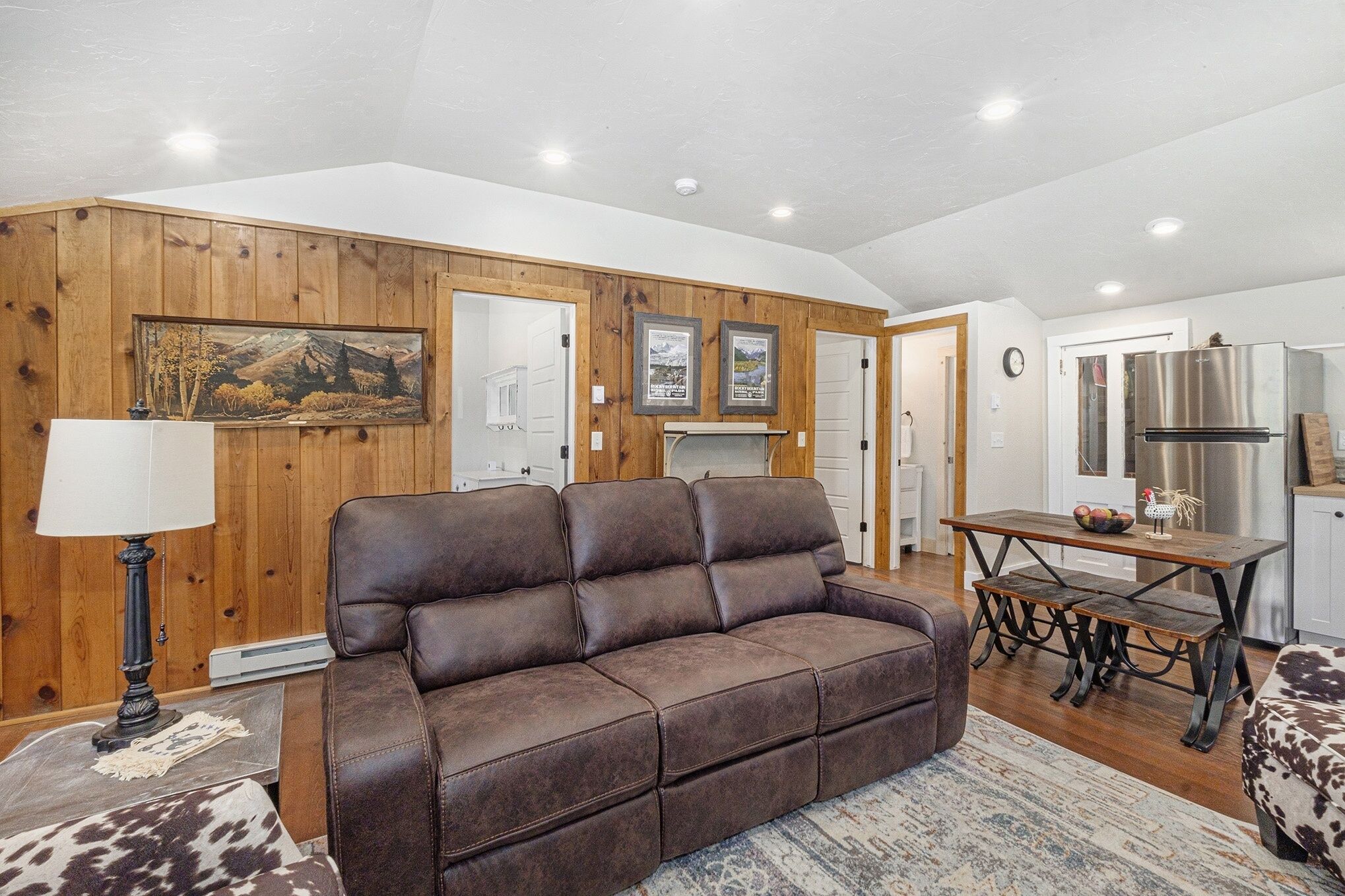 A living room with a brown leather sofa, a wooden dining table, and a kitchen area with a stainless steel refrigerator.