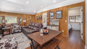 A spacious living area with a brown leather couch, a wooden dining table with a fruit bowl, and a cowhide-patterned chair. - Niwot Cabin - Stay In Rocky Mountain National Park - Estes Park - New Listing - 22-zone3298 2 Bedroom Cabin by RedAwning (Estes Park)