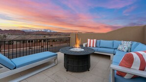 Eine Terrasse mit einem runden Feuerkorb, blauer und weißer Möbel und Blick auf die Berge im Abendrot.